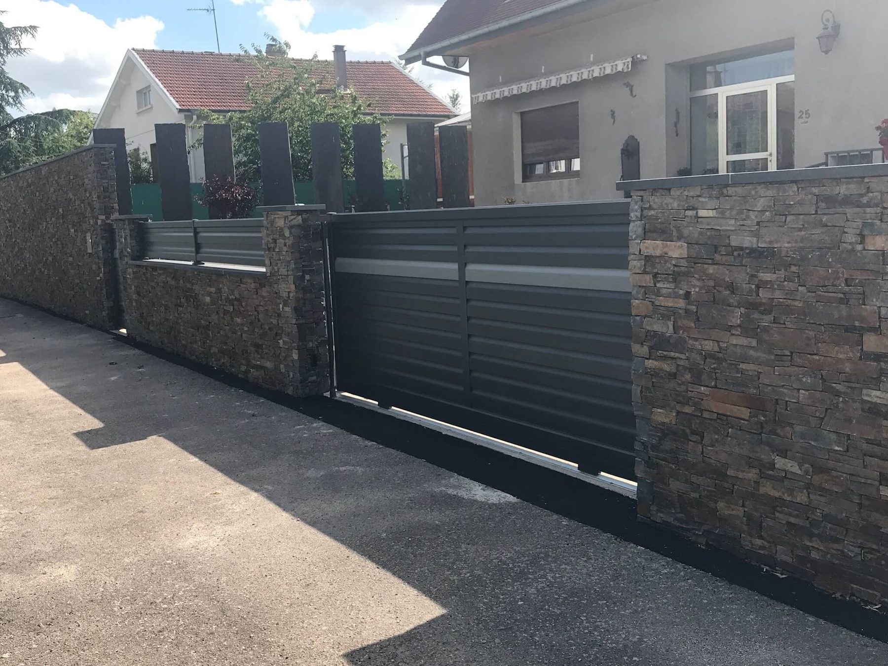 Portail coulissant gris entre deux murs en parement de pierre, ardoises dressées derrière