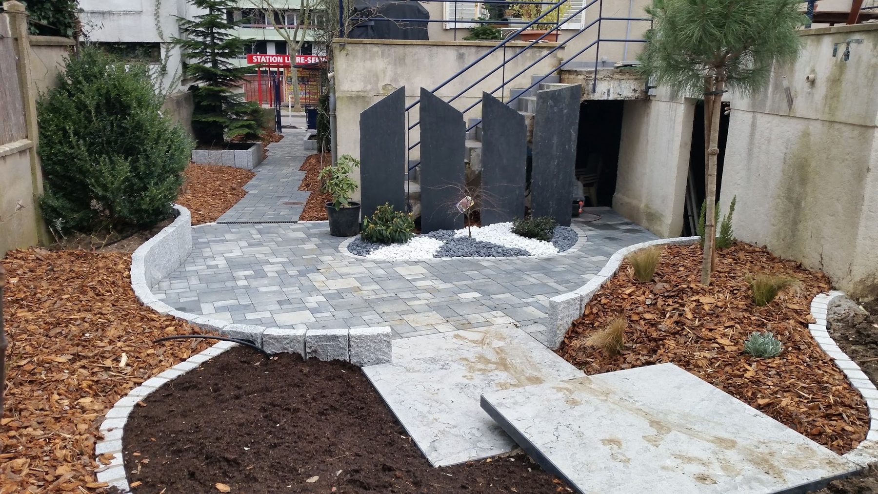 Cour pavée bordée de granit, palissade d'ardoises dressées, massifs paillés et jeune pin
